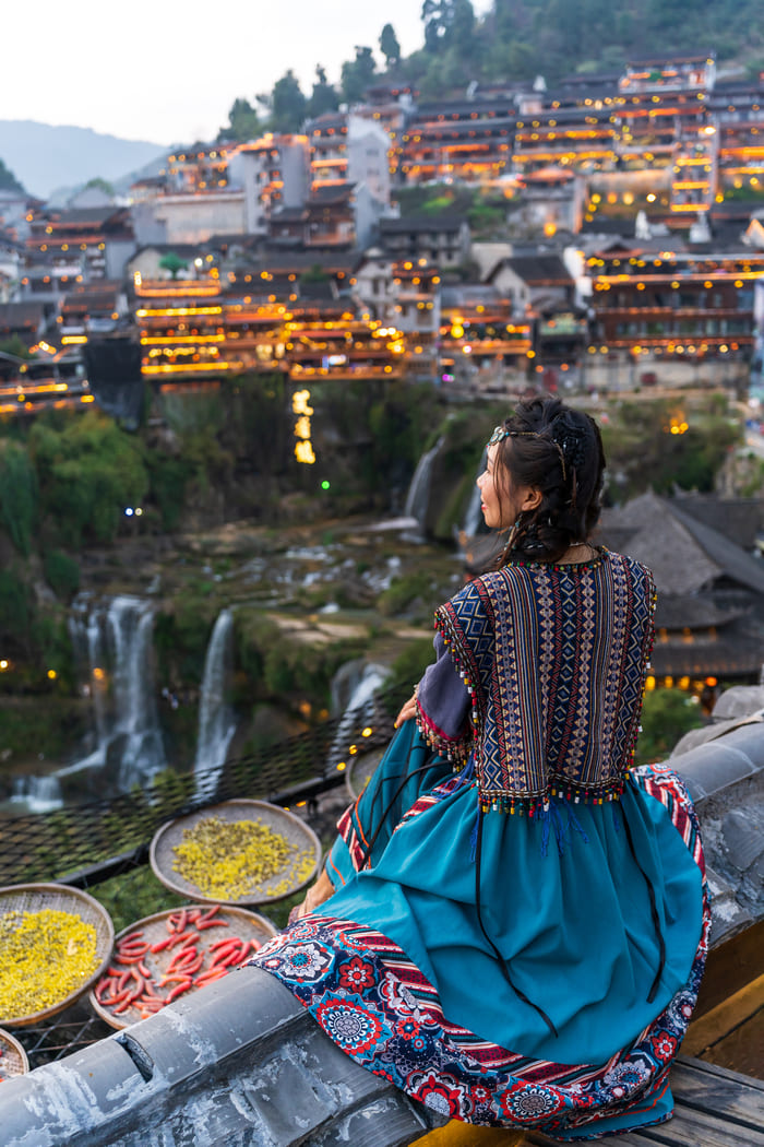 Mujer con vestimenta tradicional contemplando el paisaje de una antigua ciudad china lijiang iluminada al atardecer, rodeada de arquitectura tradicional, montañas y cascadas.