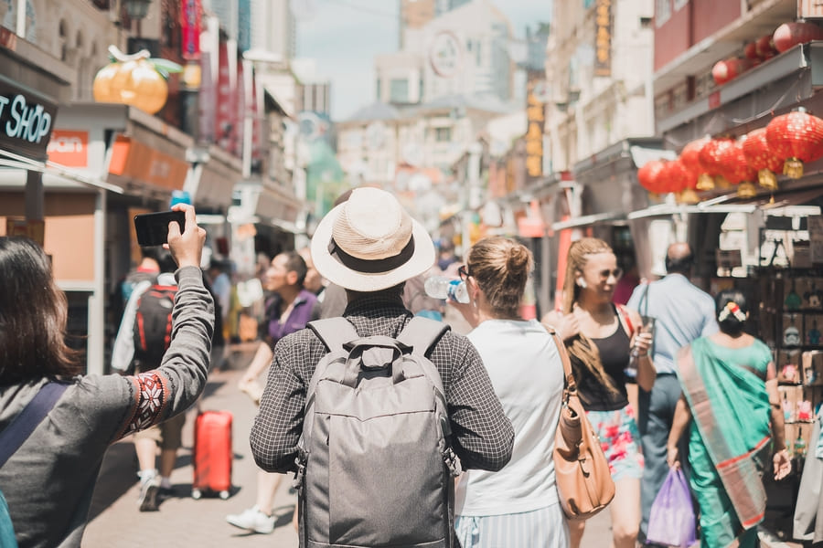 Turistas recorriendo una concurrida calle comercial de una ciudad china, rodeados de tiendas, comercios y faroles rojos tradicionales.